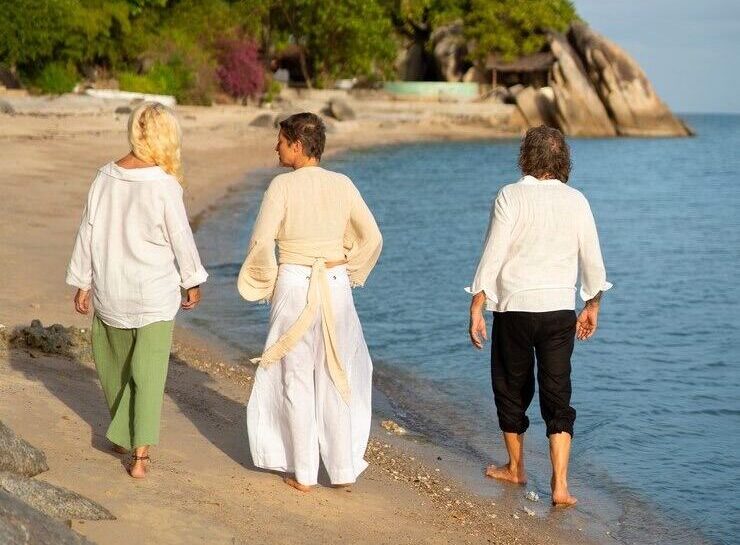 Tres personas caminando descalzas por la playa, vestidas con ropa ligera de verano, relajadas y en armonía con el entorno, reflejando bienestar y tranquilidad